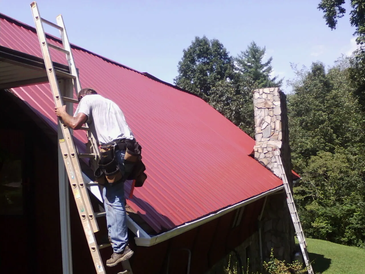 Roofer on a ladder beside a red metal roof and stone chimney — Soffit & Fascia Repair by Roofing Brothers in Ft Mitchell